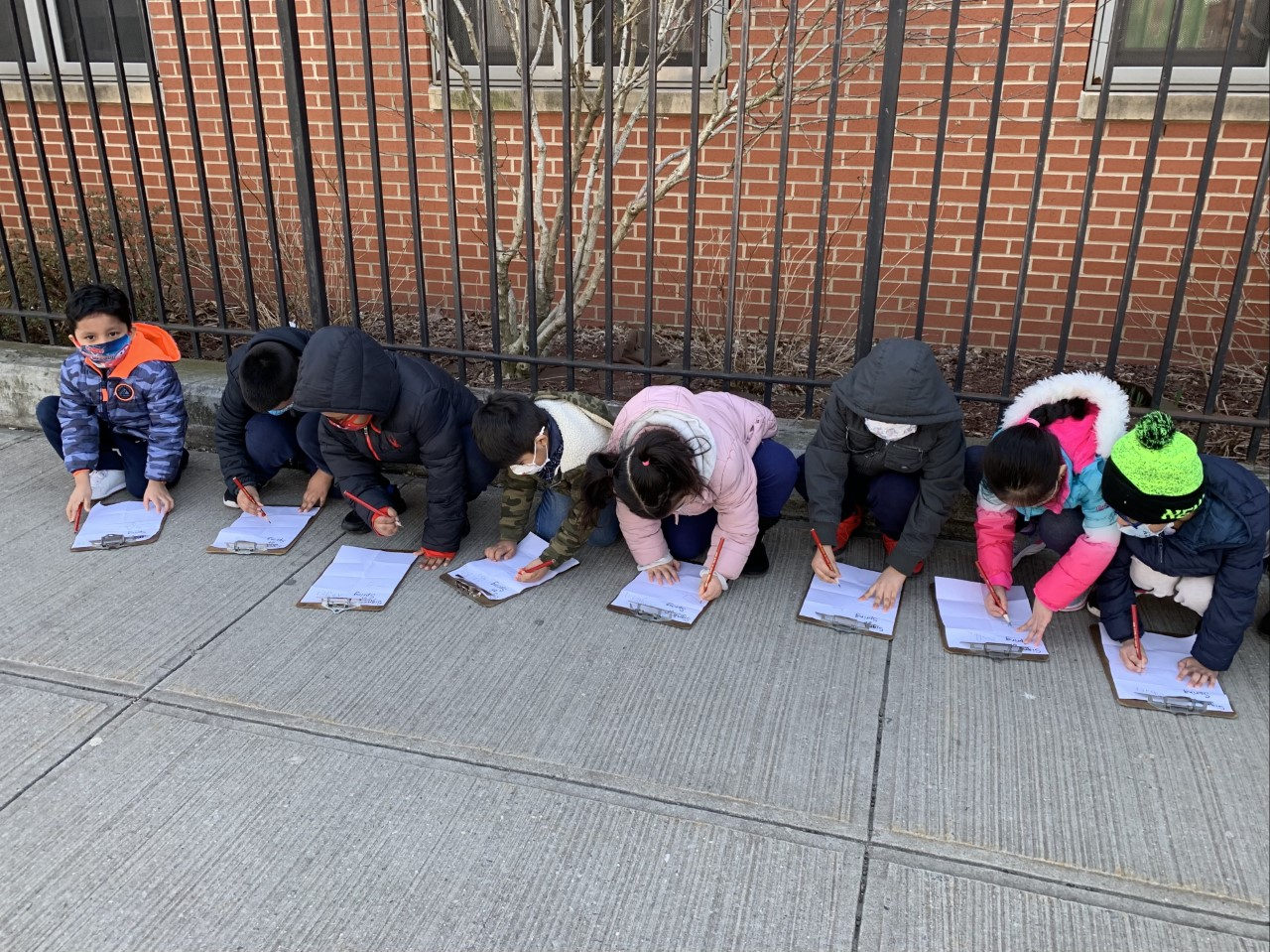 Students kneeling in a row along the fence writing on clipboards
