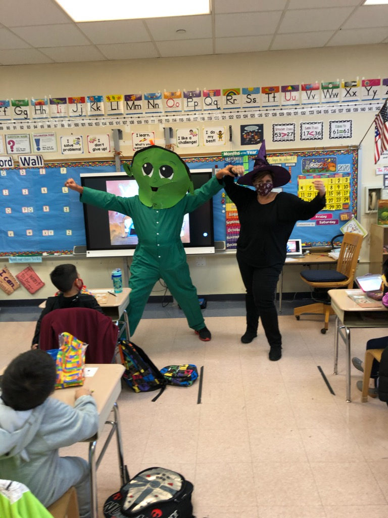 Z the green character and a teacher in a witch hat visiting a classroom