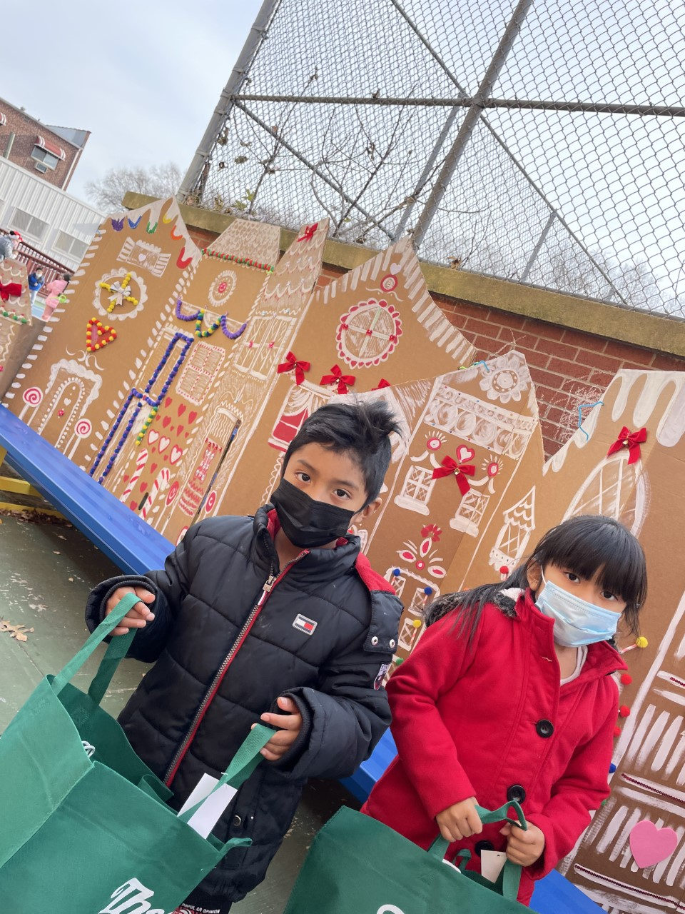 Two students with gift bags in front of a gingerbread house display