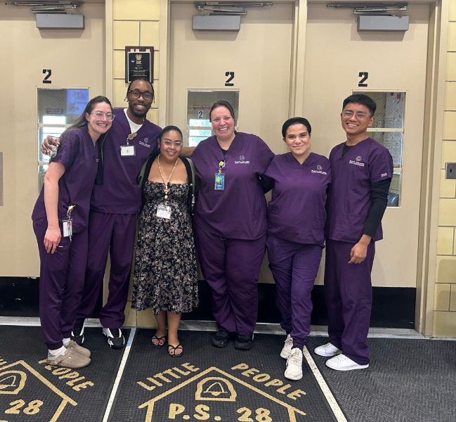Visitors in purple scrubs posing with a staff member at the school entrance