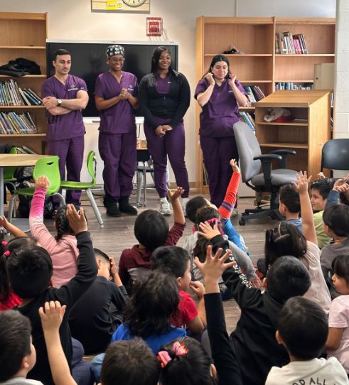 Visitors in purple scrubs speaking to students seated on the classroom floor