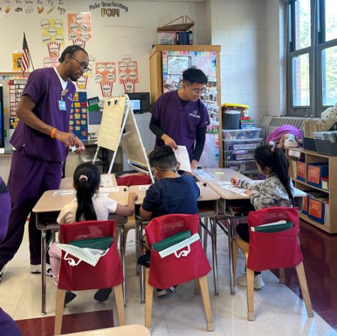 Visitors in purple scrubs talking with students at their desks