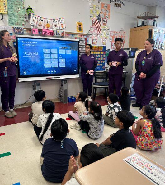 Visitors in purple scrubs presenting to students seated on the carpet