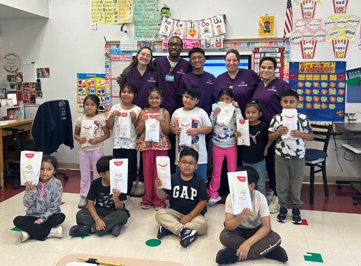 Class posing with visitors in purple scrubs, holding dental goodie bags