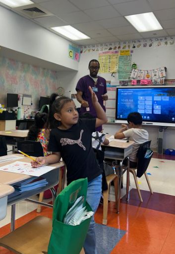 Students raising their hands as a visitor in purple scrubs presents at the smartboard