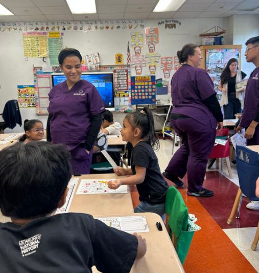 Visitors in purple scrubs working with students at their tables