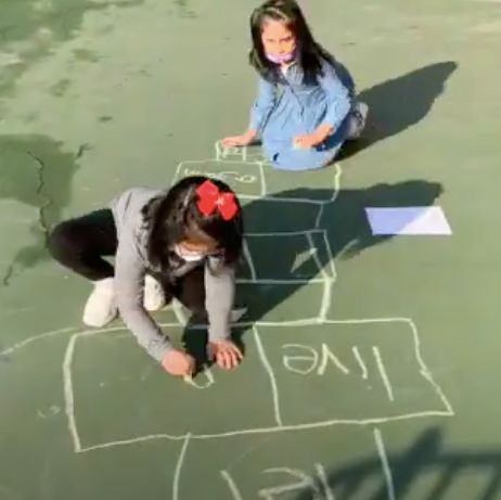 Two students drawing a chalk hopscotch board with words written in the squares