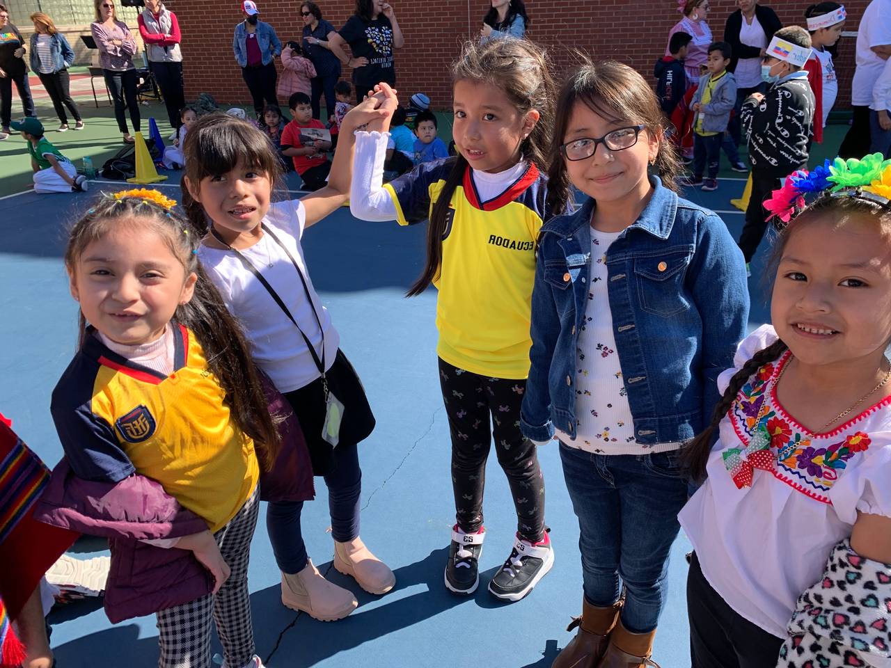 Smiling P.S. 28 students in soccer jerseys and traditional dress hold hands on the schoolyard court.