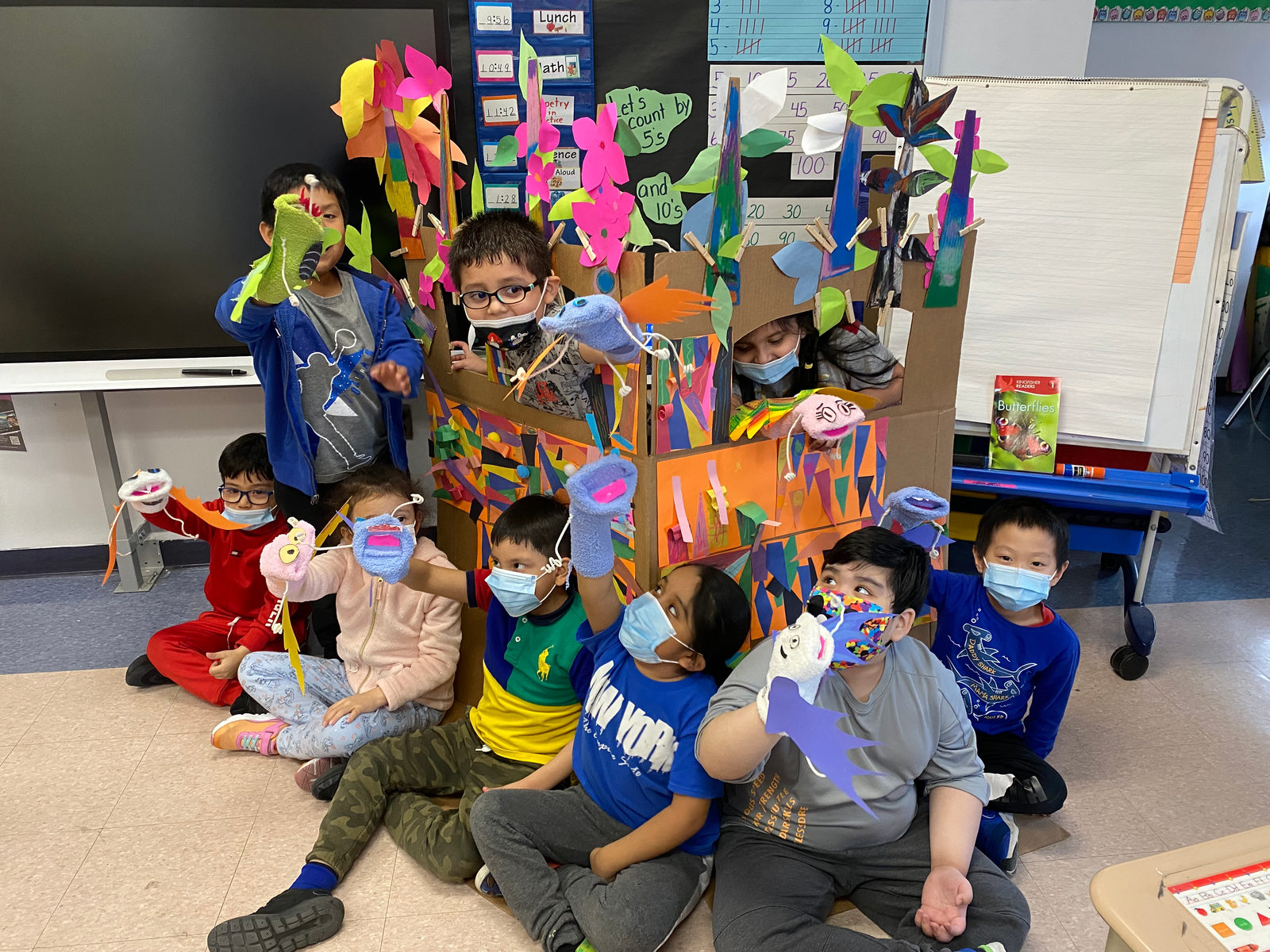 Students smile and hold up their puppets beside their class puppet stage