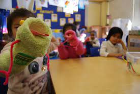 Students at a classroom table hold up the sock puppets they made