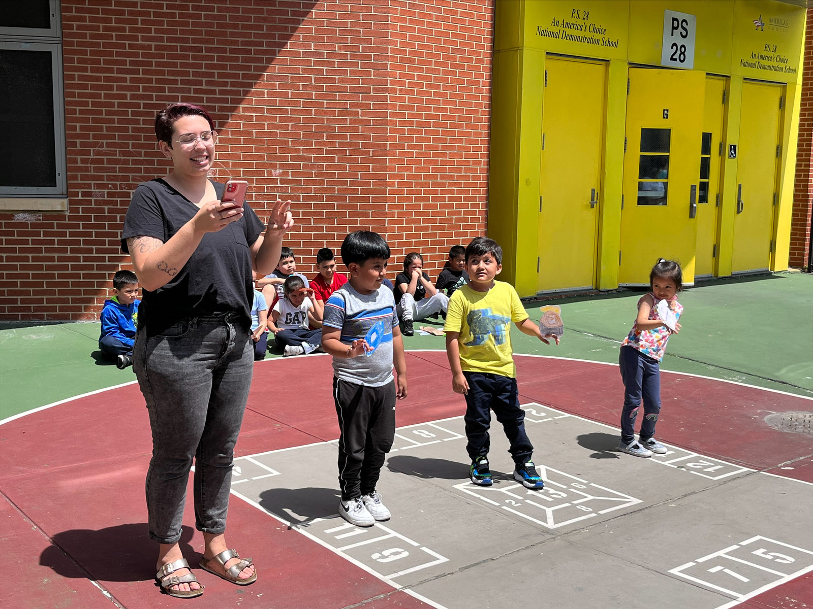 Students step forward to perform near the yellow schoolyard doors while their classmates watch