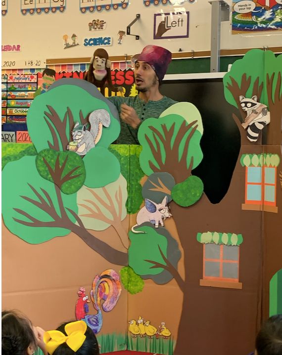 A puppeteer performs from behind a painted tree set decorated with animal puppets