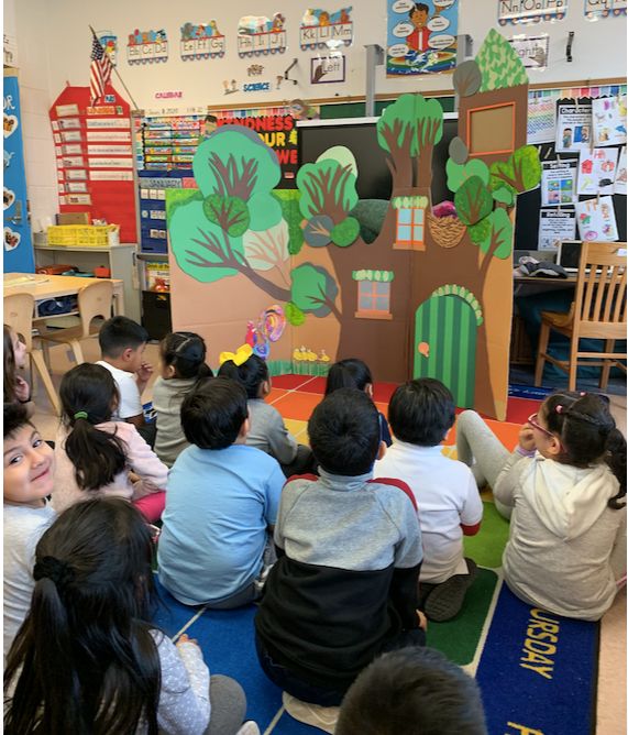 Students sit on the rug watching a puppet show performed from a painted cardboard tree set