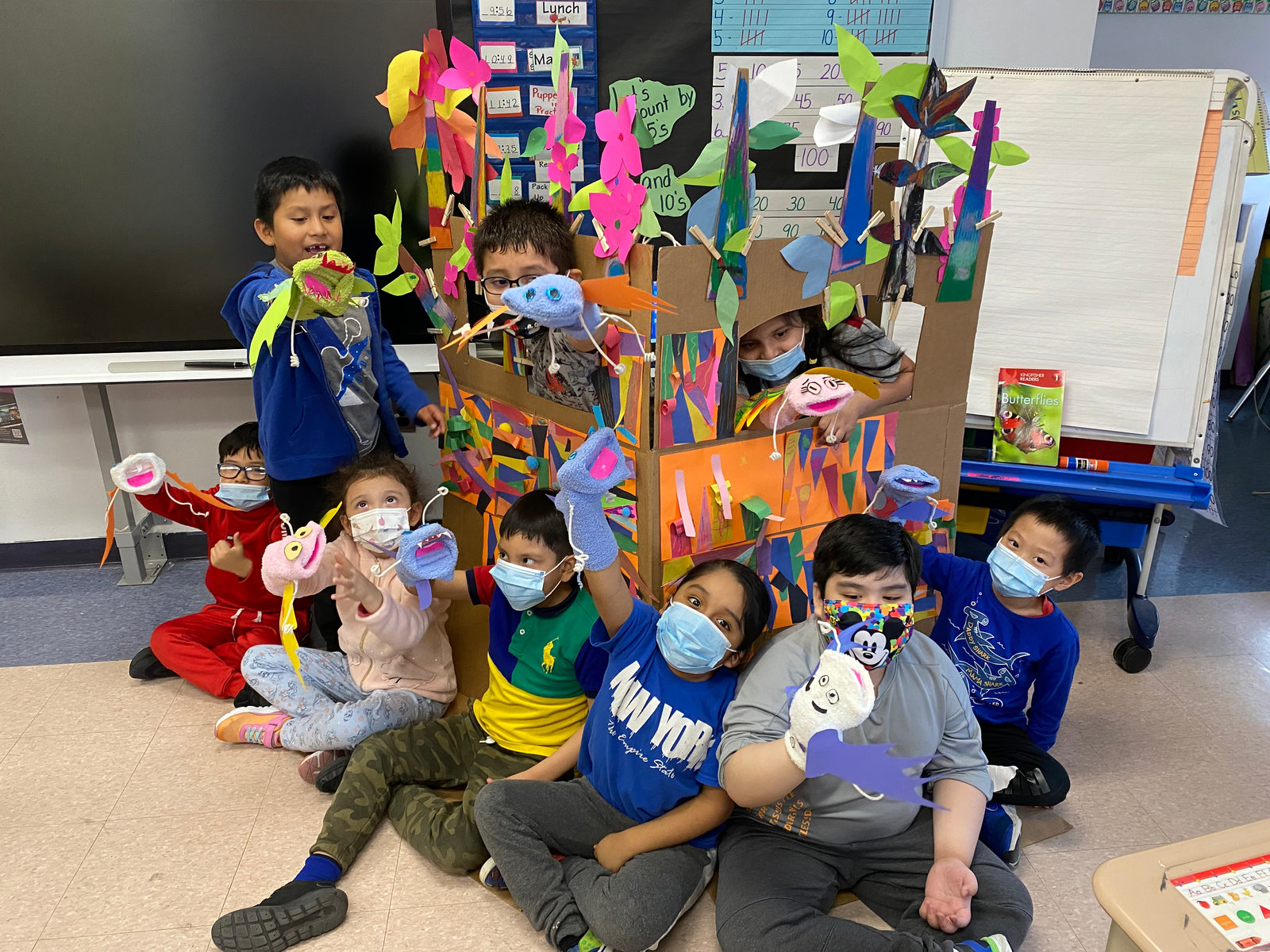 A class sits together with their puppets in front of the colorful puppet stage they built