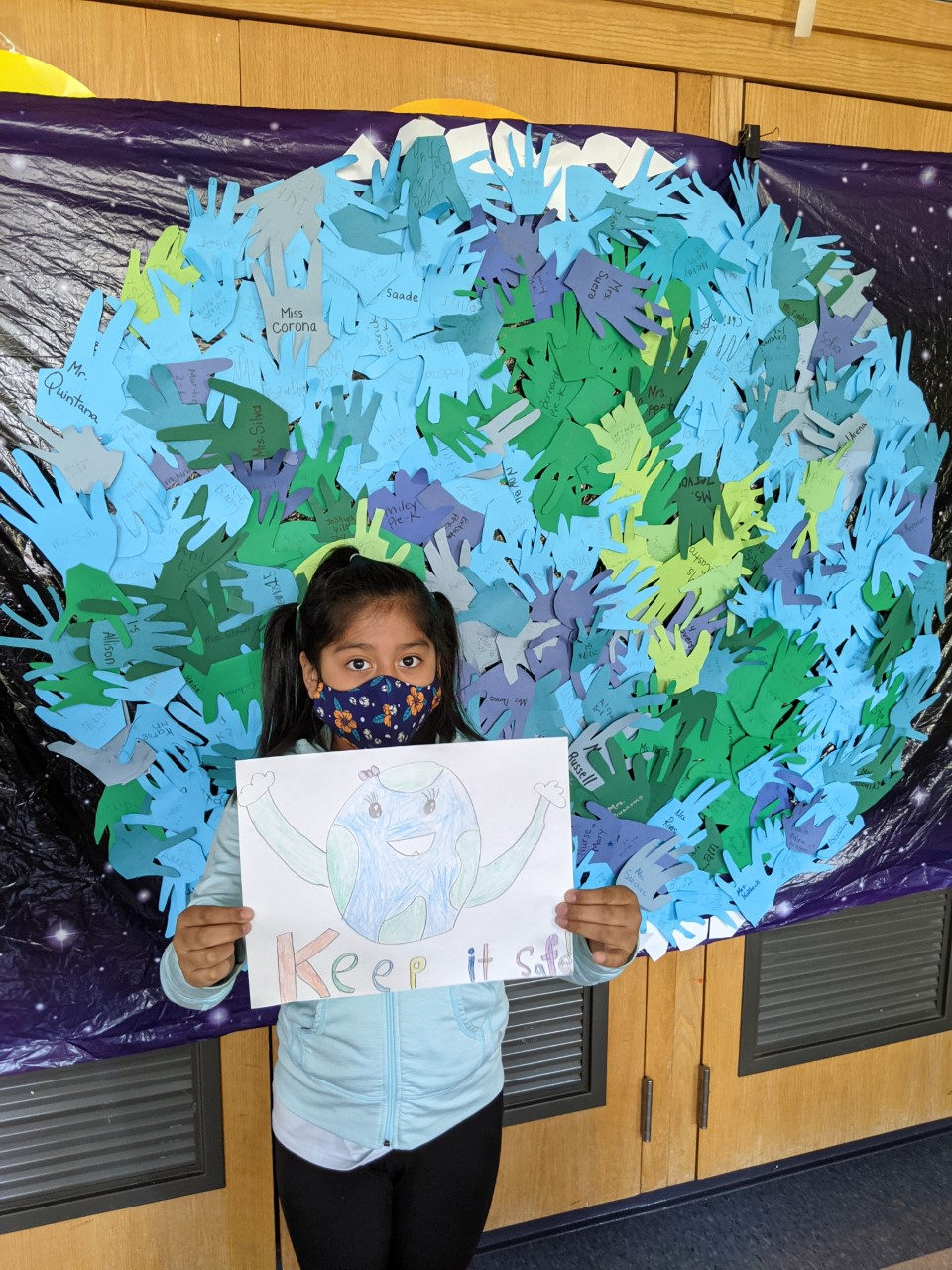 Student holding a Keep It Safe drawing in front of the hand-print Earth