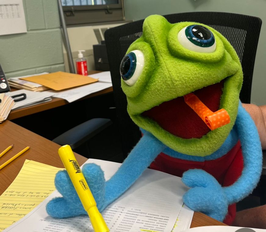 A green chameleon puppet holding a yellow highlighter at a desk