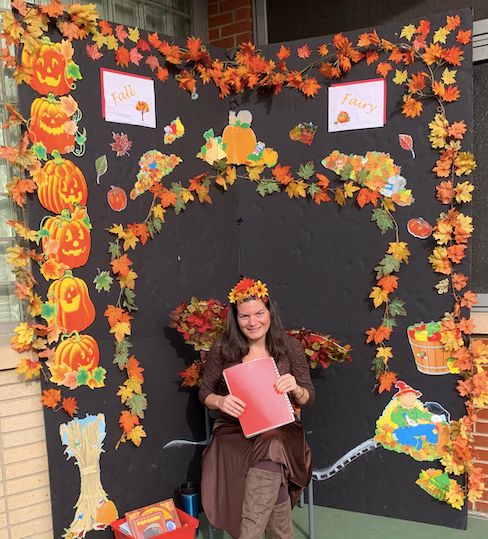 Teacher in a leaf crown in front of a fall display of pumpkins and autumn leaves