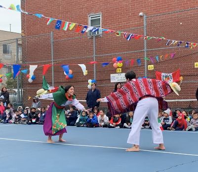 Dancers in traditional dress performing for students in the schoolyard under strings of flags